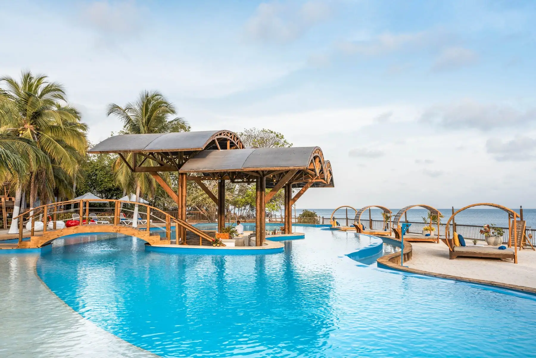 Piscina de borde curvo con puente de madera y palmeras en el hotel Isla del Encanto